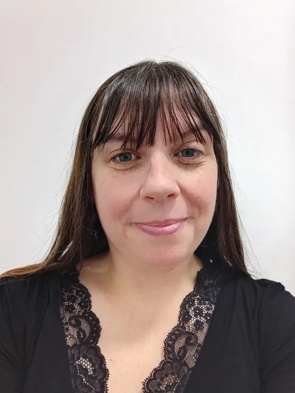 Portrait of staff member Jenifer Keenan with long dark brown hair wearing a black top with a lace neckline, facing the camera against a plain white background.